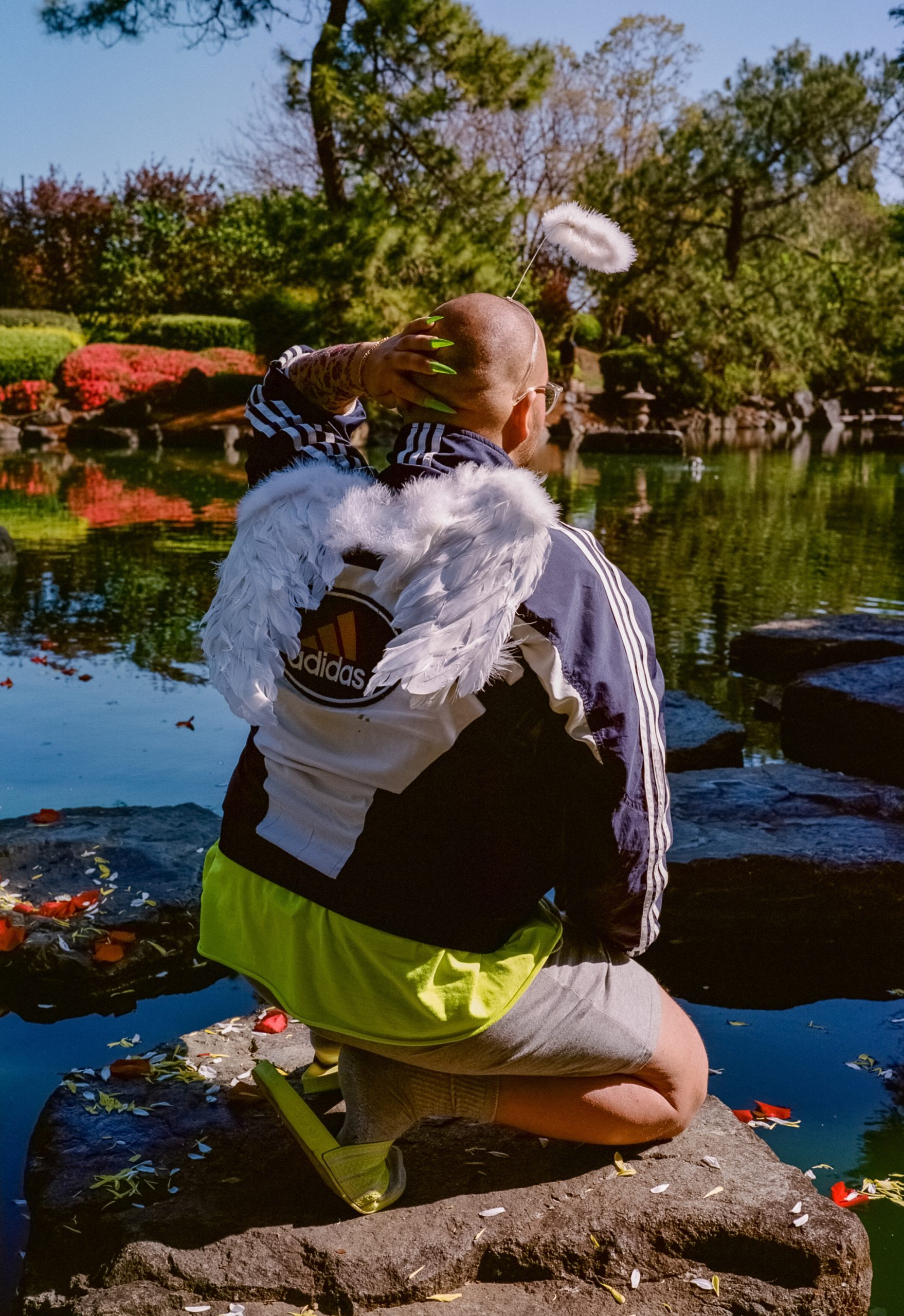 A figure is squatting on a stone with their back to the camera amidst the pond of the Auburn Botanic Japanese Gardens in this photographic portrait. Red, yellow and white flower petals line the pond and the stone. The colorful flora of the gardens can be seen in the background. The non-binary figure is wearing bright green acrylic nail clip-ons, a bright green t-shirt, bright green slides, and a navy blue Adidas wind-breaker. Feathered wings adorn their back, a toy-store halo is floating above their head, and fake tattoos are lined down their fore-arm. Their hand is raised and resting behind their head.